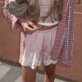 Person wearing a plaid shirt over a white top and pink shorts with lace trim against a brown wall.